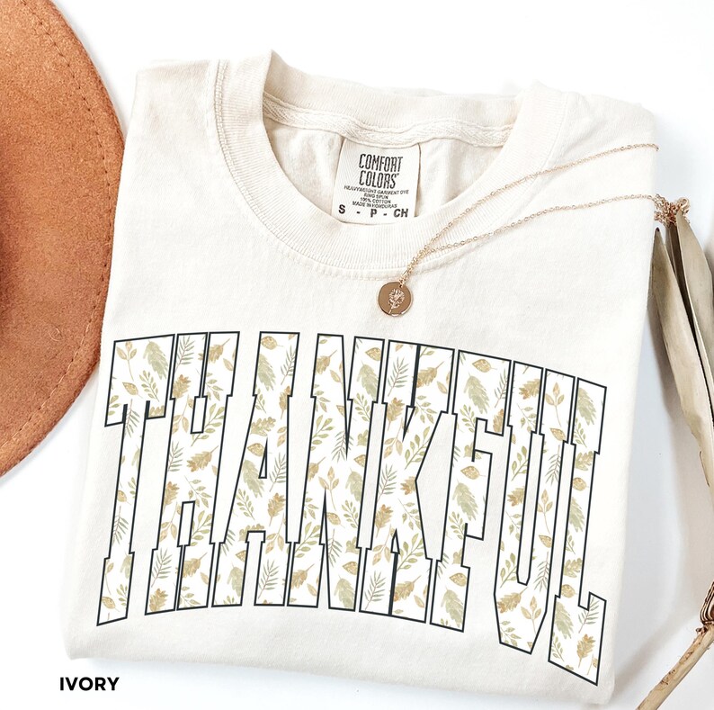May include: Ivory-colored t-shirt with the word "THANKFUL" in large, block letters filled with a floral pattern of green and gold leaves. A gold necklace with a pendant rests on the shirt. The shirt is folded.