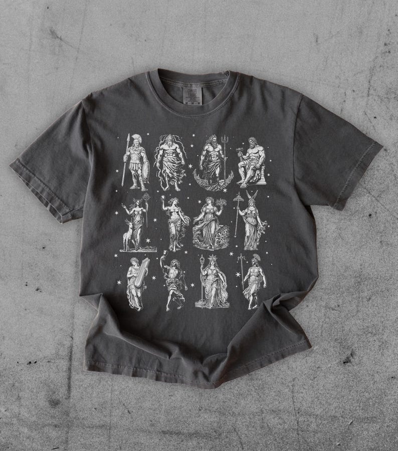 May include: Dark gray t-shirt featuring a detailed white illustration of various Greek gods and goddesses. The design includes figures like Zeus, Poseidon, and Athena, with stars scattered around the images. The shirt is made of a soft material.