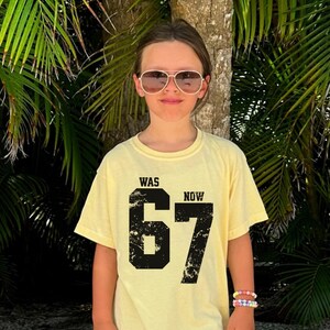 Peut inclure: Un t-shirt &agrave; manches courtes jaune p&acirc;le avec un grand imprim&eacute; graphique noir comportant le texte "WAS" et "NOW" au-dessus du nombre "67". Le nombre "67" pr&eacute;sente une texture vieillie et us&eacute;e. Le t-shirt a un col rond.