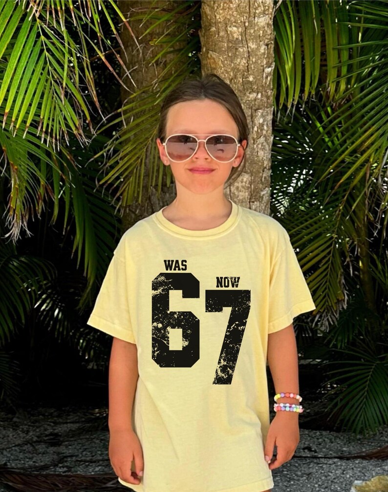 Peut inclure: Un t-shirt &agrave; manches courtes jaune p&acirc;le avec un grand imprim&eacute; graphique noir comportant le texte "WAS" et "NOW" au-dessus du nombre "67". Le nombre "67" pr&eacute;sente une texture vieillie et us&eacute;e. Le t-shirt a un col rond.