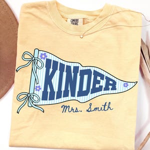 Puede incluir: Una camiseta de color amarillo mantequilla con un gráfico de un banderín a rayas azul claro y blanco con el texto KINDER en letras de bloque azul oscuro. Debajo del banderín, el texto Mrs. Smith está impreso en una fuente cursiva azul oscuro.