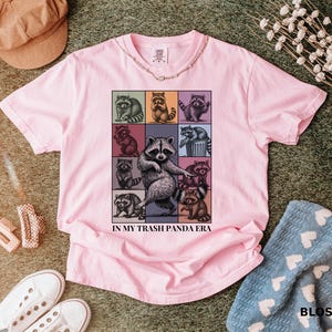 May include: Pink t-shirt featuring a grid of raccoon illustrations in various poses, with the text "IN MY TRASH PANDA ERA". The shirt is surrounded by accessories, including a corduroy hat and white sneakers.
