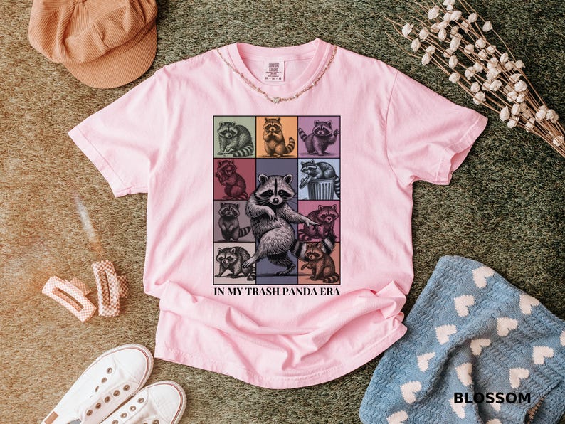 May include: Pink t-shirt featuring a grid of raccoon illustrations in various poses, with the text "IN MY TRASH PANDA ERA". The shirt is surrounded by accessories, including a corduroy hat and white sneakers.