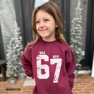 Peut inclure: Un sweat-shirt &agrave; col rond et manches longues de couleur bordeaux avec une impression graphique blanche vieillie sur le devant. Le motif affiche le mot WAS au-dessus du nombre 67 et le mot NOW &agrave; droite du nombre dans une police sans empattement.