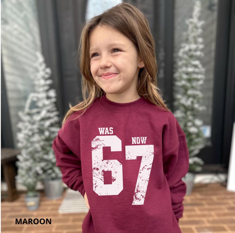 Peut inclure: Un sweat-shirt &agrave; col rond et manches longues de couleur bordeaux avec une impression graphique blanche vieillie sur le devant. Le motif affiche le mot WAS au-dessus du nombre 67 et le mot NOW &agrave; droite du nombre dans une police sans empattement.