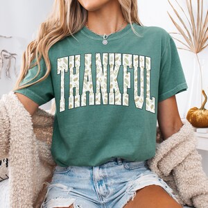 May include: Light green t-shirt with the word "THANKFUL" in large, white, block letters. The letters are filled with a beige leaf pattern. The shirt is short-sleeved and has a relaxed fit. The text "LIGHT GREEN" is at the bottom of the image.