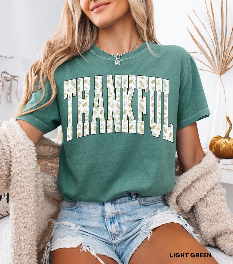 May include: Light green t-shirt with the word "THANKFUL" in large, white, block letters. The letters are filled with a beige leaf pattern. The shirt is short-sleeved and has a relaxed fit. The text "LIGHT GREEN" is at the bottom of the image.
