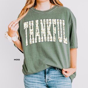 May include: Moss green t-shirt with the word "THANKFUL" in large, white, block letters filled with a leaf pattern. The shirt is paired with blue jeans. The word "MOSS" is printed in black at the bottom left.