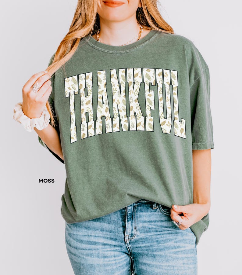 May include: Moss green t-shirt with the word "THANKFUL" in large, white, block letters filled with a leaf pattern. The shirt is paired with blue jeans. The word "MOSS" is printed in black at the bottom left.
