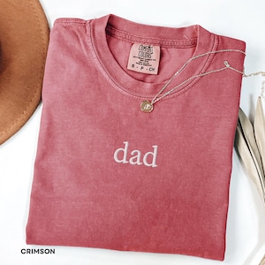May include: Crimson-colored t-shirt with the word "dad" embroidered in white. A silver necklace with a pendant rests on the shirt. The shirt has a crew neck and a tag that reads "Comfort Colors."