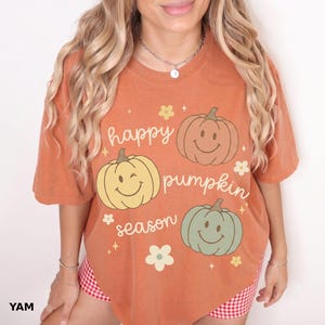 Puede incluir: Una camiseta de manga corta color naranja quemado con tres ilustraciones de calabazas con caras sonrientes en tonos marrón, amarillo y verde salvia. El texto "happy pumpkin season" está impreso en cursiva blanca, con pequeñas flores y estrellas amarillas.