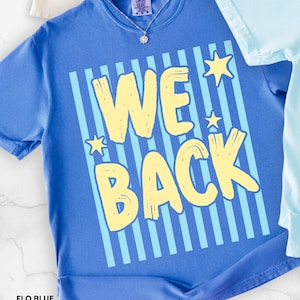 Puede incluir: Una camiseta de manga corta color azul bígaro con un diseño gráfico de rayas verticales azul claro y el texto WE BACK en letras grandes de color amarillo pálido con efecto de pincelada. Tres pequeñas estrellas amarillas rodean el texto.
