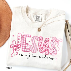 May include: Ivory-colored t-shirt with the words "JESUS is my love story" in pink, patterned letters. The letters have gingham, striped, and cherry patterns. A gold necklace with a pendant and a brown hat are also visible.