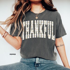 May include: A dark gray t-shirt with the word "THANKFUL" in large, white, block letters. The letters are filled with a pattern of green and tan leaves. The word "PEPPER" is printed in small, white letters below the shirt.