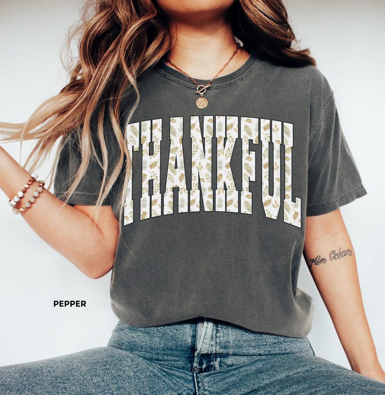 May include: A dark gray t-shirt with the word "THANKFUL" in large, white, block letters. The letters are filled with a pattern of green and tan leaves. The word "PEPPER" is printed in small, white letters below the shirt.