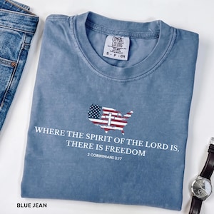 May include: A blue jean-colored t-shirt with the text "WHERE THE SPIRIT OF THE LORD IS, THERE IS FREEDOM" and "2 CORINTHIANS 3:17". The shirt features a graphic of the United States map with a cross and American flag design. The shirt is from Comfort Colors.