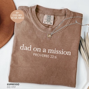 Puede incluir: Una camiseta marrón con el texto "dad on a mission" y "PROVERBS 22:6" en blanco. La camiseta está hecha de un material suave y cómodo y está disponible en más de 10 colores.