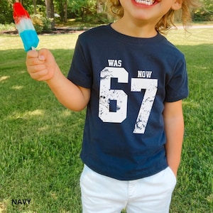 Peut inclure: Un t-shirt bleu marine &agrave; manches courtes avec le texte blanc WAS NOW et le grand chiffre 67 dans un style de police us&eacute; et vieilli. Le t-shirt est associ&eacute; &agrave; un pantalon blanc, et une glace rouge, blanche et bleue est tenue en l'air.