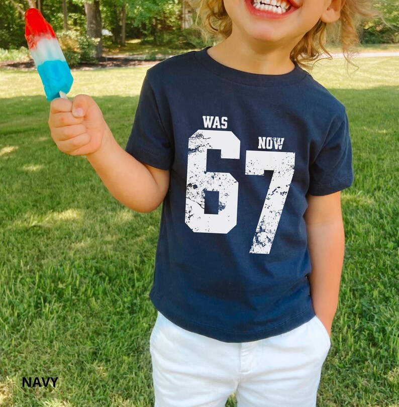 Peut inclure: Un t-shirt bleu marine &agrave; manches courtes avec le texte blanc WAS NOW et le grand chiffre 67 dans un style de police us&eacute; et vieilli. Le t-shirt est associ&eacute; &agrave; un pantalon blanc, et une glace rouge, blanche et bleue est tenue en l'air.