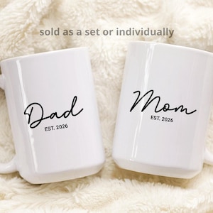 Puede incluir: Dos tazas de cerámica blancas con texto en escritura negra que dice "Dad" y "Mom" con "EST. 2026" debajo. Las tazas tienen asas grandes y están sobre un fondo blanco suave.