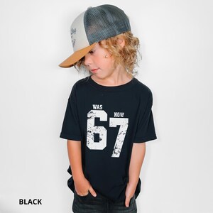 Peut inclure: Un t-shirt noir &agrave; manches courtes avec le texte blanc WAS et NOW au-dessus des grands chiffres 67 &agrave; l'aspect vieilli. Il est associ&eacute; &agrave; une casquette de type trucker avec une visi&egrave;re beige, un panneau avant blanc et un filet gris fonc&eacute;.
