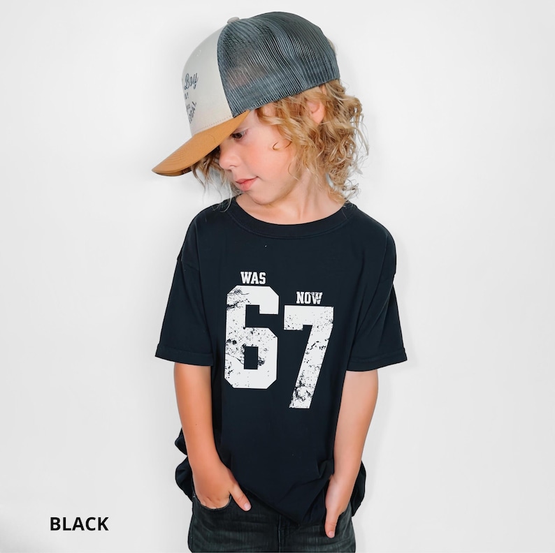 Peut inclure: Un t-shirt noir &agrave; manches courtes avec le texte blanc WAS et NOW au-dessus des grands chiffres 67 &agrave; l'aspect vieilli. Il est associ&eacute; &agrave; une casquette de type trucker avec une visi&egrave;re beige, un panneau avant blanc et un filet gris fonc&eacute;.