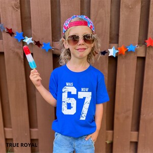 Peut inclure: Un t-shirt bleu vif avec le texte blanc WAS 67 NOW 67 dans une police us&eacute;e. Le t-shirt est port&eacute; avec une casquette &agrave; motifs multicolores, des lunettes de soleil aux verres bruns et un jean bleu, tout en tenant une glace rouge, blanche et bleue.