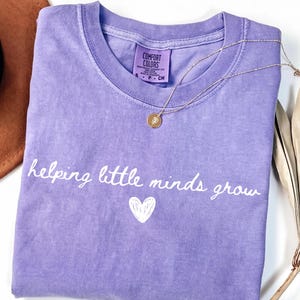 Peut inclure: Un t-shirt violet à manches courtes avec un col rond et une étiquette Comfort Colors. Le devant présente le texte blanc "helping little minds grow" en écriture cursive au-dessus d'un petit cœur blanc dessiné à la main. Un collier doré est posé dessus.