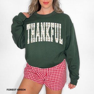 May include: Forest green sweatshirt with the word "THANKFUL" in white block letters filled with a beige floral pattern. The person is wearing red and white checkered shorts. The sweatshirt is a long-sleeved crew neck.