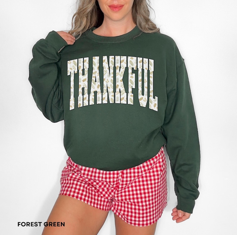 May include: Forest green sweatshirt with the word "THANKFUL" in white block letters filled with a beige floral pattern. The person is wearing red and white checkered shorts. The sweatshirt is a long-sleeved crew neck.
