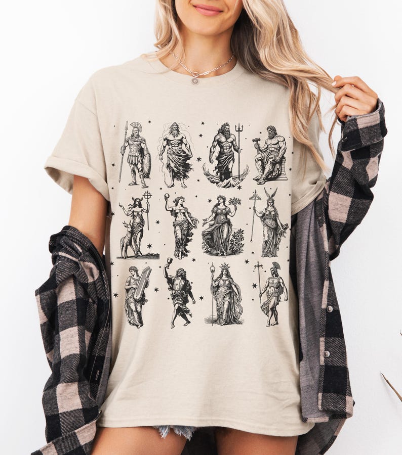 May include: Beige t-shirt featuring a black and white illustration of various Greek gods and goddesses. The graphic includes figures like Zeus, Poseidon, and Athena, with stars scattered around the images. The shirt has short sleeves and a crew neck.