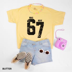Peut inclure: Un t-shirt &agrave; manches courtes jaune p&acirc;le avec le texte noir vieilli WAS 67 NOW, un short en jean bleu clair &agrave; taille crois&eacute;e, des lunettes de soleil blanches en forme de fleur, un collier de perles pastel et un appareil photo instantan&eacute; rose.