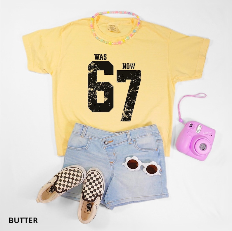 Peut inclure: Un t-shirt &agrave; manches courtes jaune p&acirc;le avec le texte noir vieilli WAS 67 NOW, un short en jean bleu clair &agrave; taille crois&eacute;e, des lunettes de soleil blanches en forme de fleur, un collier de perles pastel et un appareil photo instantan&eacute; rose.
