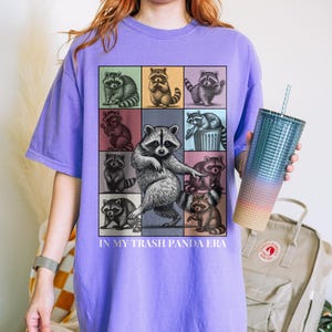 May include: Violet-colored t-shirt featuring a grid of raccoon illustrations with the text "IN MY TRASH PANDA ERA." The design includes raccoons in various poses and colors. A gradient tumbler with a straw is held in the hand.