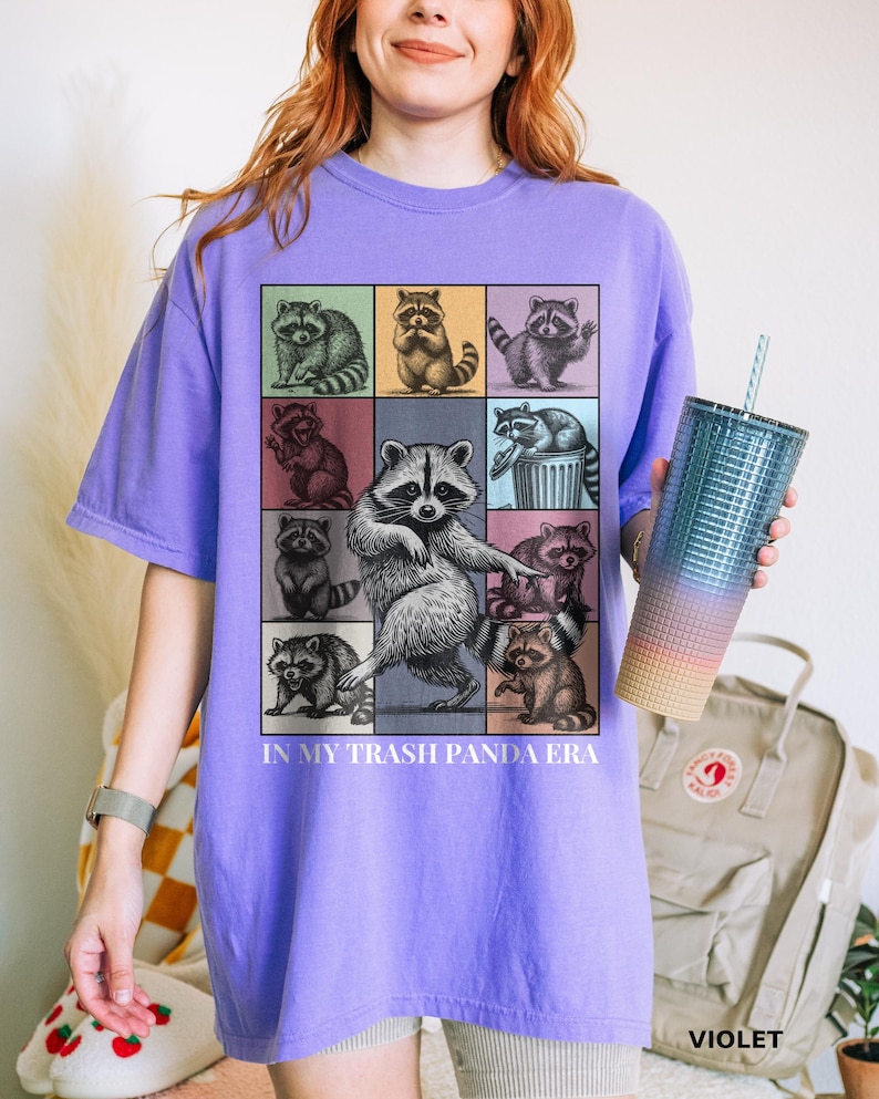 May include: Violet-colored t-shirt featuring a grid of raccoon illustrations with the text "IN MY TRASH PANDA ERA." The design includes raccoons in various poses and colors. A gradient tumbler with a straw is held in the hand.