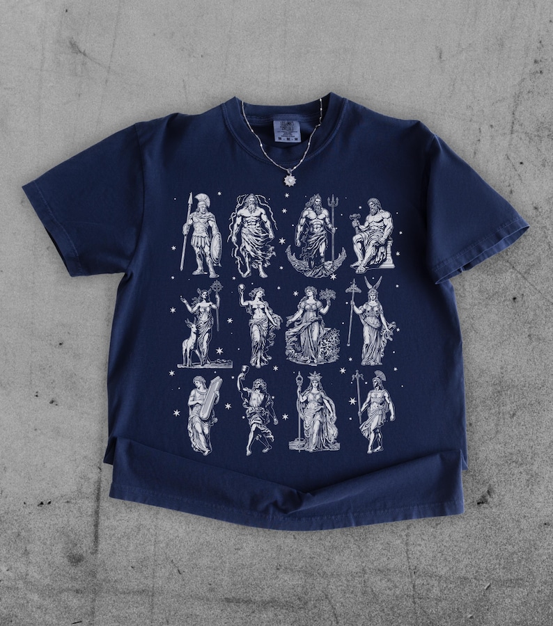 May include: Navy blue t-shirt featuring a white illustration of Greek gods and goddesses. The graphic includes figures like Zeus and Poseidon. The shirt has a classic crew neck and short sleeves, and is made of a soft material.
