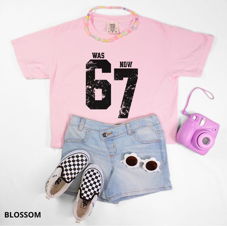 Peut inclure: Un t-shirt rose clair avec le texte noir vieilli WAS NOW 67, un short en jean bleu clair, des baskets &agrave; enfiler &agrave; damier noir et blanc, un collier de perles pastel, des lunettes de soleil en forme de fleur et un appareil photo instantan&eacute; violet.