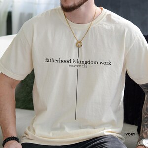 May include: Ivory-colored t-shirt with the text "fatherhood is kingdom work" and "PROVERBS 22:6" printed in black. A vertical black line extends from the text. The shirt is a short-sleeved, crew neck style.