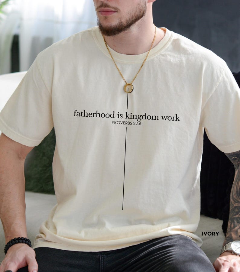 May include: Ivory-colored t-shirt with the text "fatherhood is kingdom work" and "PROVERBS 22:6" printed in black. A vertical black line extends from the text. The shirt is a short-sleeved, crew neck style.