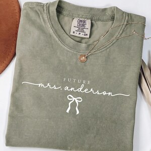 Puede incluir: Una camiseta de color verde caqui con cuello redondo y etiqueta de la marca Comfort Colors. El frente presenta texto blanco que dice FUTURE mrs. anderson con un pequeño gráfico de lazo blanco debajo. Un collar dorado descansa sobre la tela.