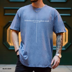 May include: Blue jean-colored t-shirt with the text "fatherhood is kingdom work" and the Bible verse Proverbs 22:6 printed in white. The shirt has a simple cross design and is made of soft material.