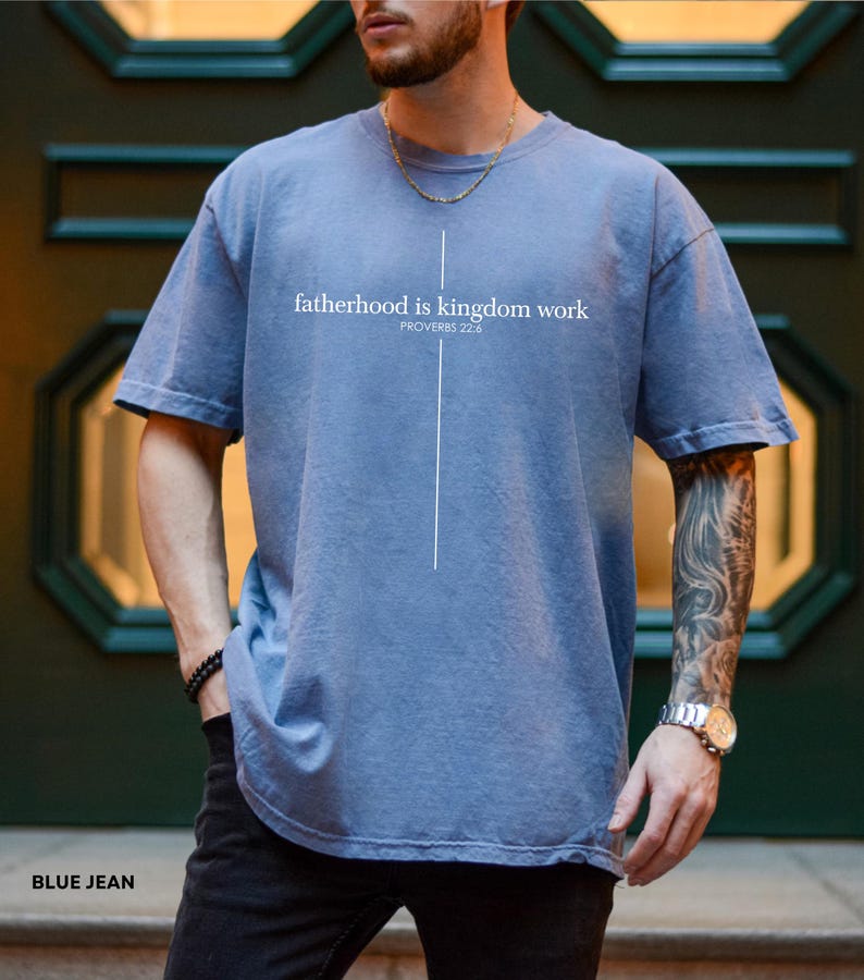 May include: Blue jean-colored t-shirt with the text "fatherhood is kingdom work" and the Bible verse Proverbs 22:6 printed in white. The shirt has a simple cross design and is made of soft material.