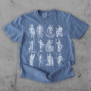 May include: A blue t-shirt featuring a white graphic of various Greek gods and goddesses. The figures are depicted in a classical style, with detailed illustrations. The shirt is made of a soft material and has a crew neck.