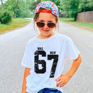 Peut inclure: Un t-shirt blanc &agrave; manches courtes avec le texte noir WAS et NOW au-dessus d'un grand chiffre 67 noir &agrave; l'aspect vieilli. Il est associ&eacute; &agrave; une casquette de baseball tie-dye rouge, blanche et bleue et &agrave; des lunettes de soleil teint&eacute;es marron.