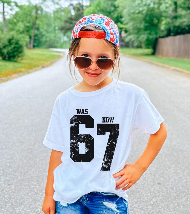 Peut inclure: Un t-shirt blanc &agrave; manches courtes avec le texte noir WAS et NOW au-dessus d'un grand chiffre 67 noir &agrave; l'aspect vieilli. Il est associ&eacute; &agrave; une casquette de baseball tie-dye rouge, blanche et bleue et &agrave; des lunettes de soleil teint&eacute;es marron.