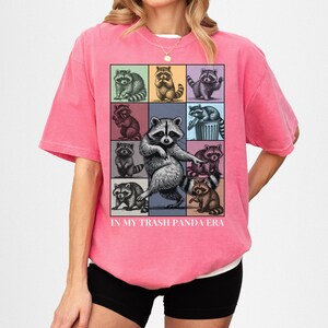 May include: A pink t-shirt featuring a grid of raccoon illustrations in various poses and colors. The phrase "IN MY TRASH PANDA ERA" is printed below the grid. The shirt is worn by a person.