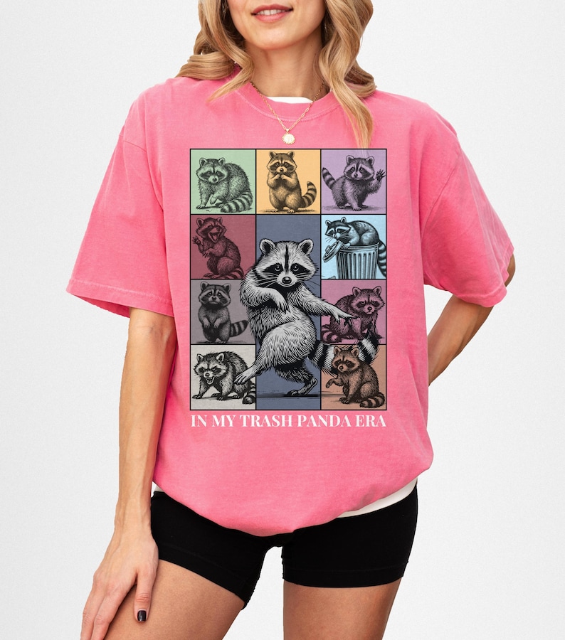 May include: A pink t-shirt featuring a grid of raccoon illustrations in various poses and colors. The phrase "IN MY TRASH PANDA ERA" is printed below the grid. The shirt is worn by a person.