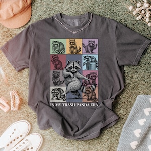 May include: A gray t-shirt featuring a grid of cartoon raccoons in various poses, with one large raccoon in the center. The text "IN MY TRASH PANDA ERA" is printed below the grid. The shirt is displayed on a textured surface.