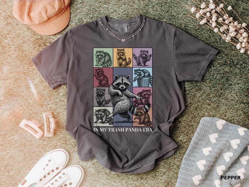May include: A gray t-shirt featuring a grid of cartoon raccoons in various poses, with one large raccoon in the center. The text "IN MY TRASH PANDA ERA" is printed below the grid. The shirt is displayed on a textured surface.