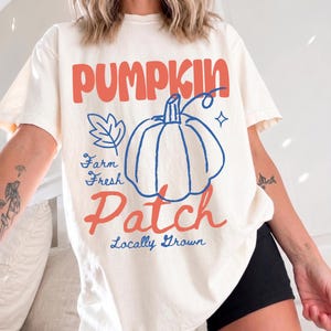 Puede incluir: Una camiseta de manga corta color marfil con un diseño gráfico que incluye el texto PUMPKIN en naranja, el contorno de una calabaza azul, una hoja azul y el texto Farm Fresh Patch Locally Grown en letras azules y naranjas.